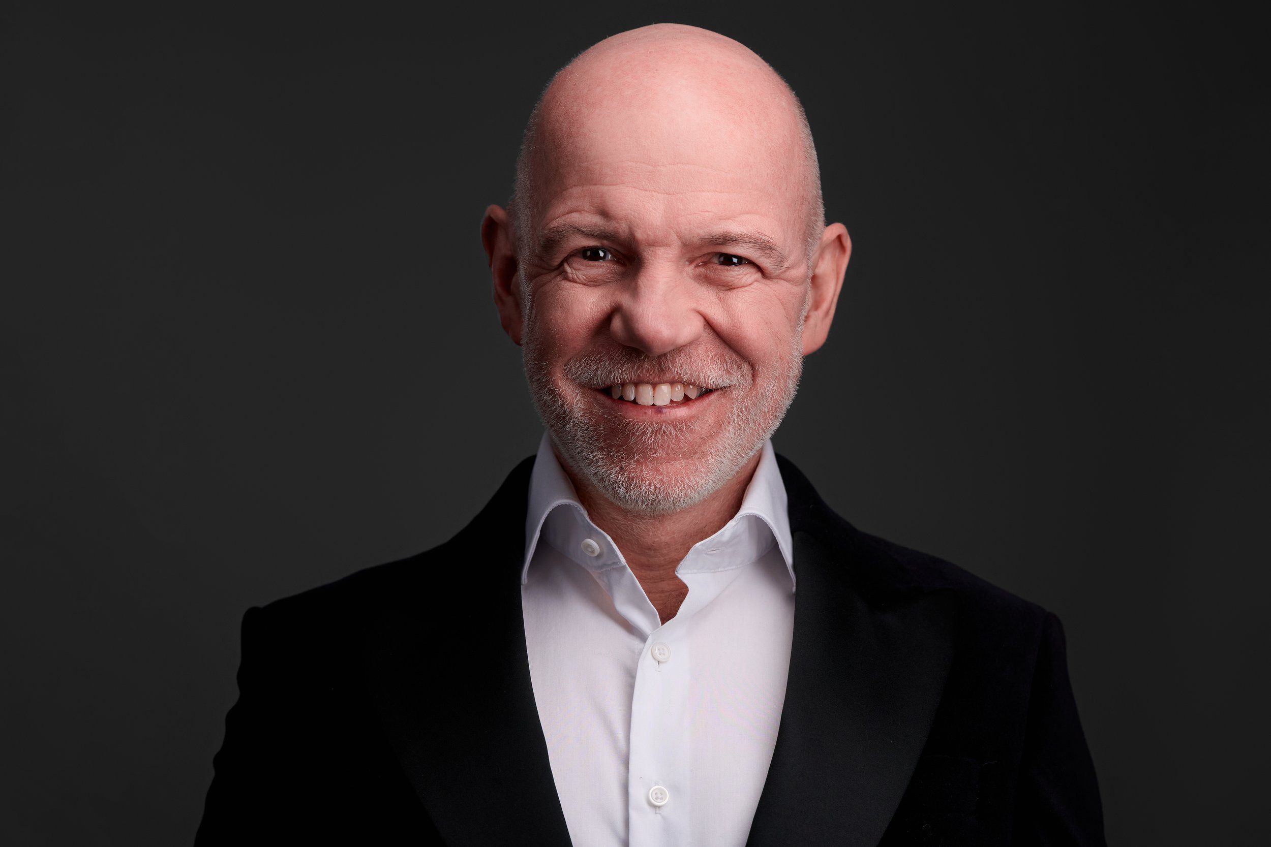 Headshot von Dietmar Wunder: freundliches Lächeln, dunkles Sakko und weißes Hemd vor dunklem Hintergrund, aufgenommen im Kölner Studio
