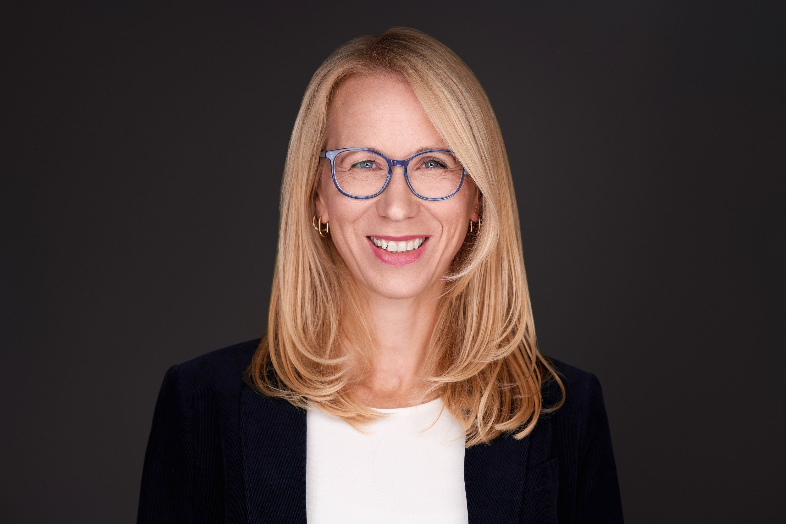 Headshot von Katrin: lächelnde Frau mit blonden Haaren und blauer Brille vor schwarzem Hintergrund